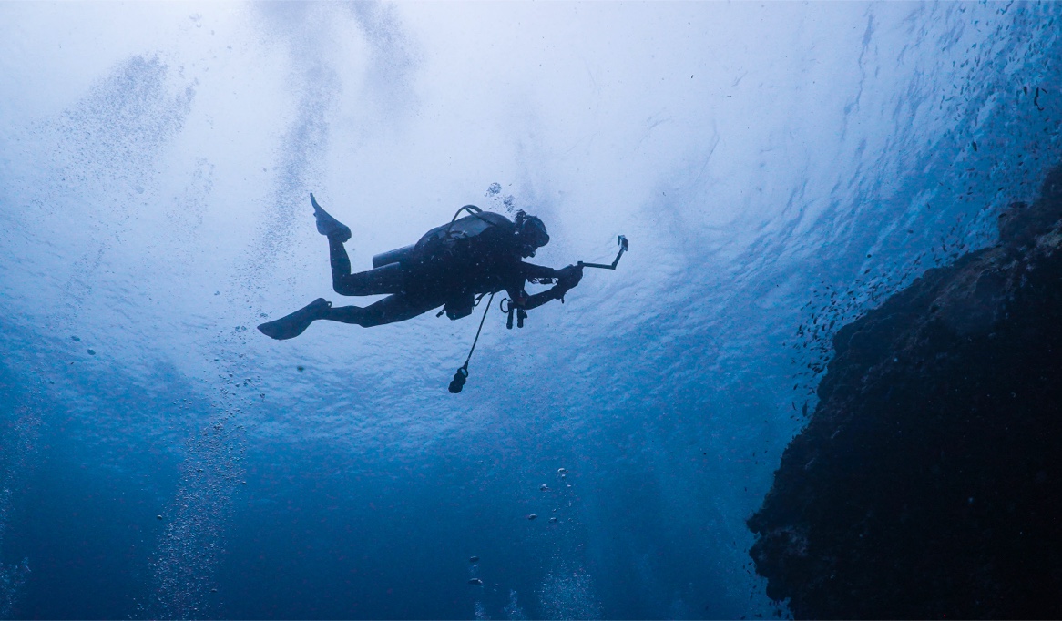 Diver silhouetted against the surface filming near a rocky wall