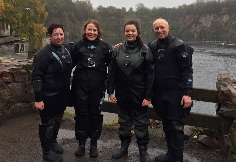 Four divers in drysuits posing beside a quarry lake