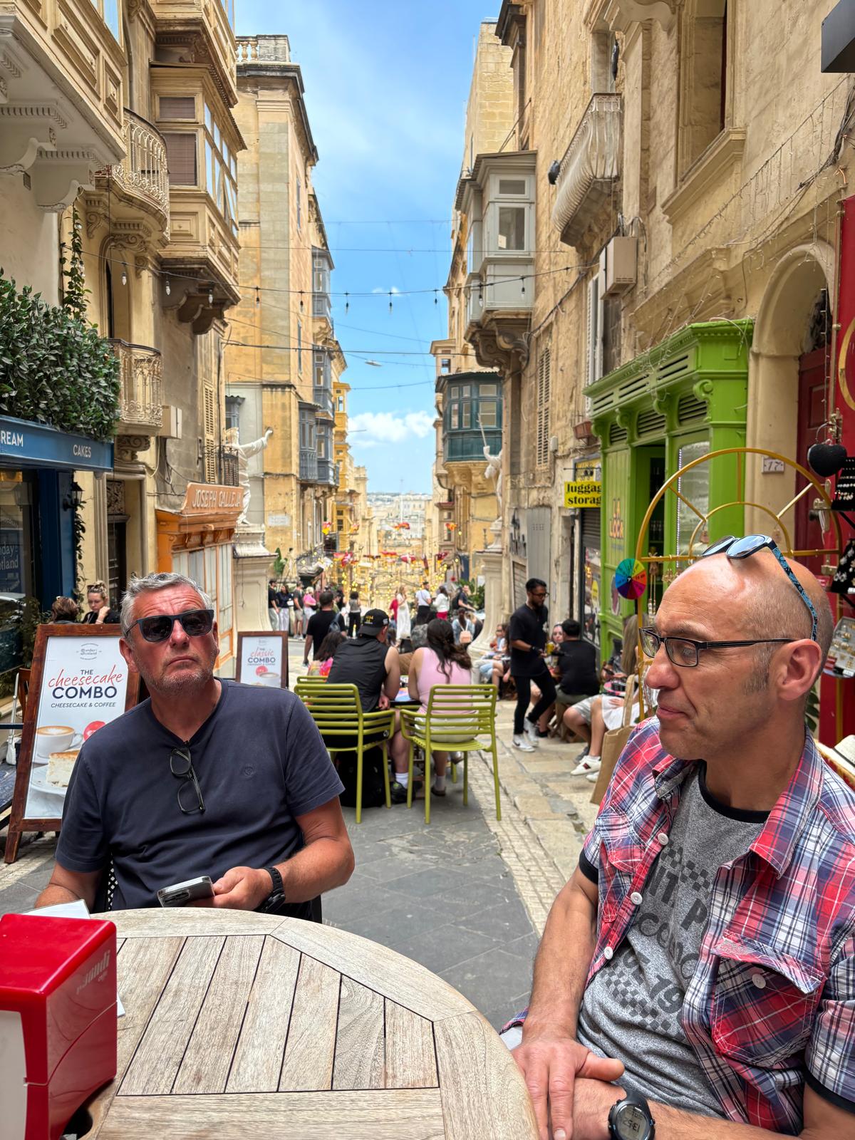 Two men seated at a café table on a narrow historic street