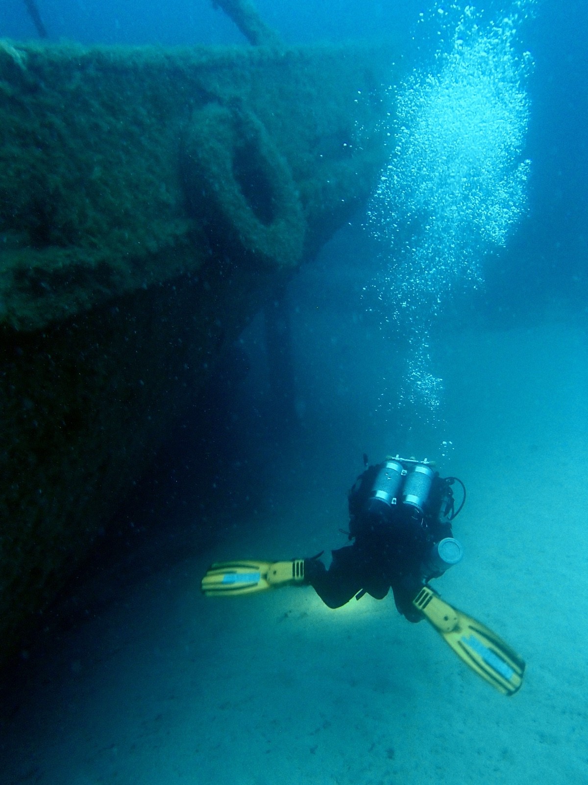 Diver with twin cylinders swimming beneath the bow of a large wreck