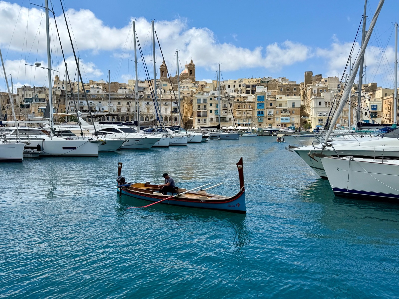 Small painted rowing boat named Malta on a harbour, with yachts and a hillside town behind