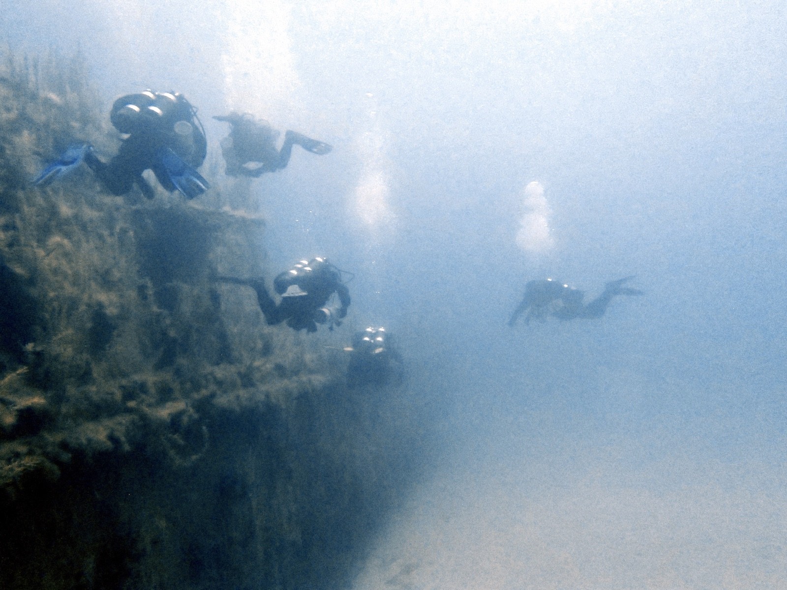 Several tec divers swimming along the encrusted side of a large wreck