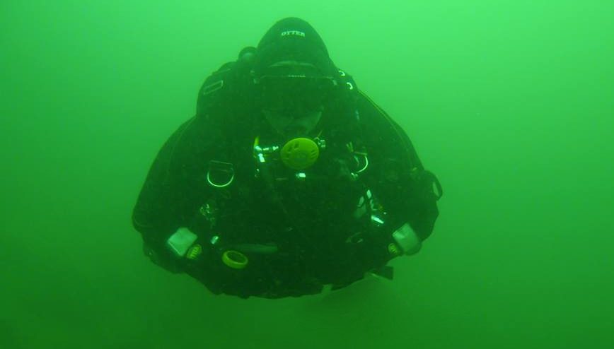 Diver in a drysuit and hood viewed from above in green water